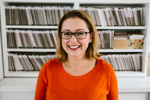 A smiling white woman in her early 40s, shoulder-length light brown hair, wearing glasses, silver necklace and a bright orange jumper.